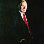 Oil painting by Josonia Palaitis depicting Justice Michael Kirby standing at turning his head to the viewer in a semi-formal pose with a red tie and a black background which envelops his suit so that it blends seamlessly and accentuates his face