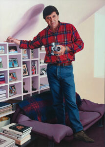 An oil painting by Josonia Palaitis depicting tv personality Ray Martin standing on a couch with a camera in his hand wearing a red an blue checked shirt with his hand resting on top of a white bookshelf with books, photos and a couple of gold tv logie awards on the shelves